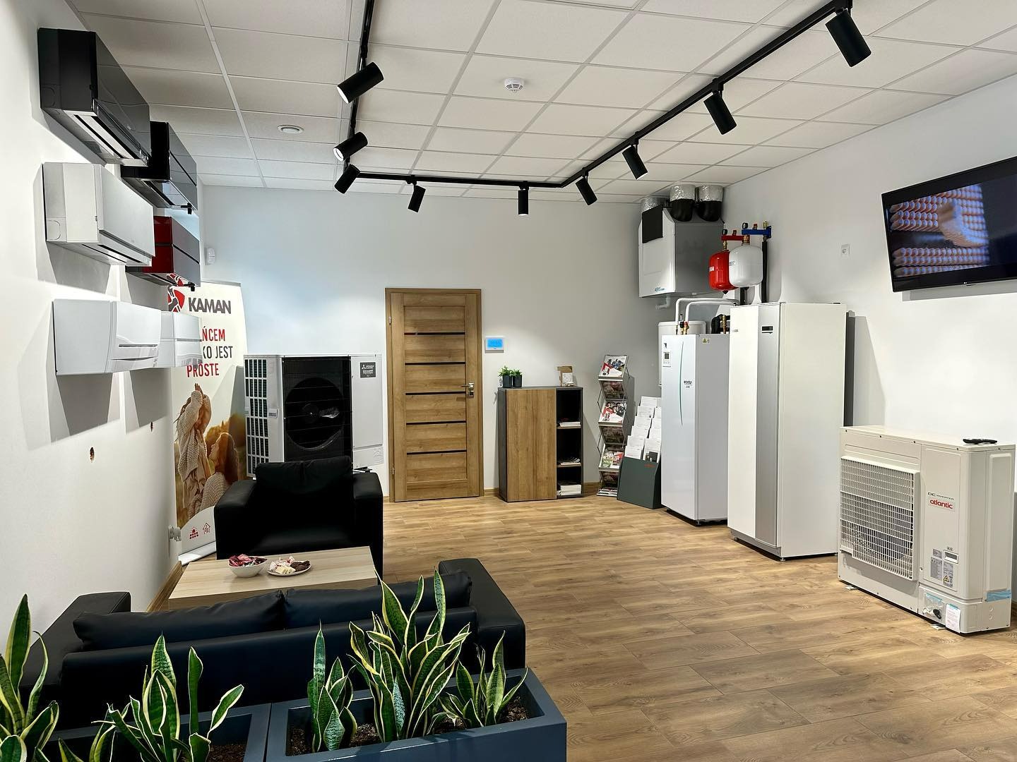 Salon ekspozycyjny z różnymi modelami urządzeń grzewczych i klimatyzacyjnych, w tym wiszące klimatyzatory, stojące jednostki i zbiornik buforowy, oświetlony reflektorami szynowymi, z sofą...