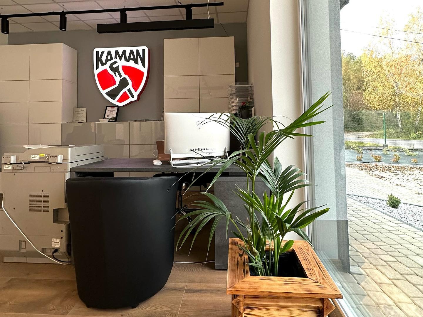 Wnętrze biura firmy instalacyjnej z logo 'KAMAN' przedstawiającym rękę trzymającą młotek na tle tarczy, widok na biurko z komputerem, drukarkę i roślinę doniczkową w drewnianej skrzynce przy oknie.