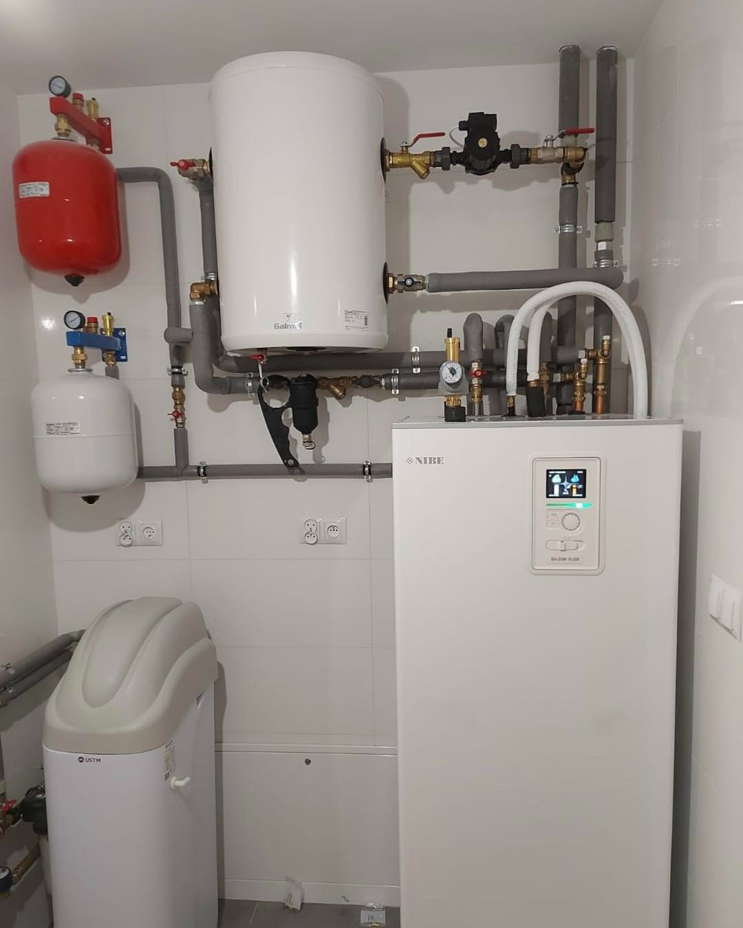 Biała kotłownia z zbiornikiem buforowym, naczyniami przeponowymi, filtrem, zmiękczaczem wody i automatyką, rury w izolacji, płytki na ścianach.