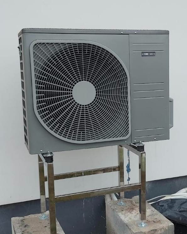 Szara jednostka zewnętrzna urządzenia grzewczego NIBE AMS10 na metalowym stelażu, zamontowana na betonowych fundamentach przy białej ścianie budynku.