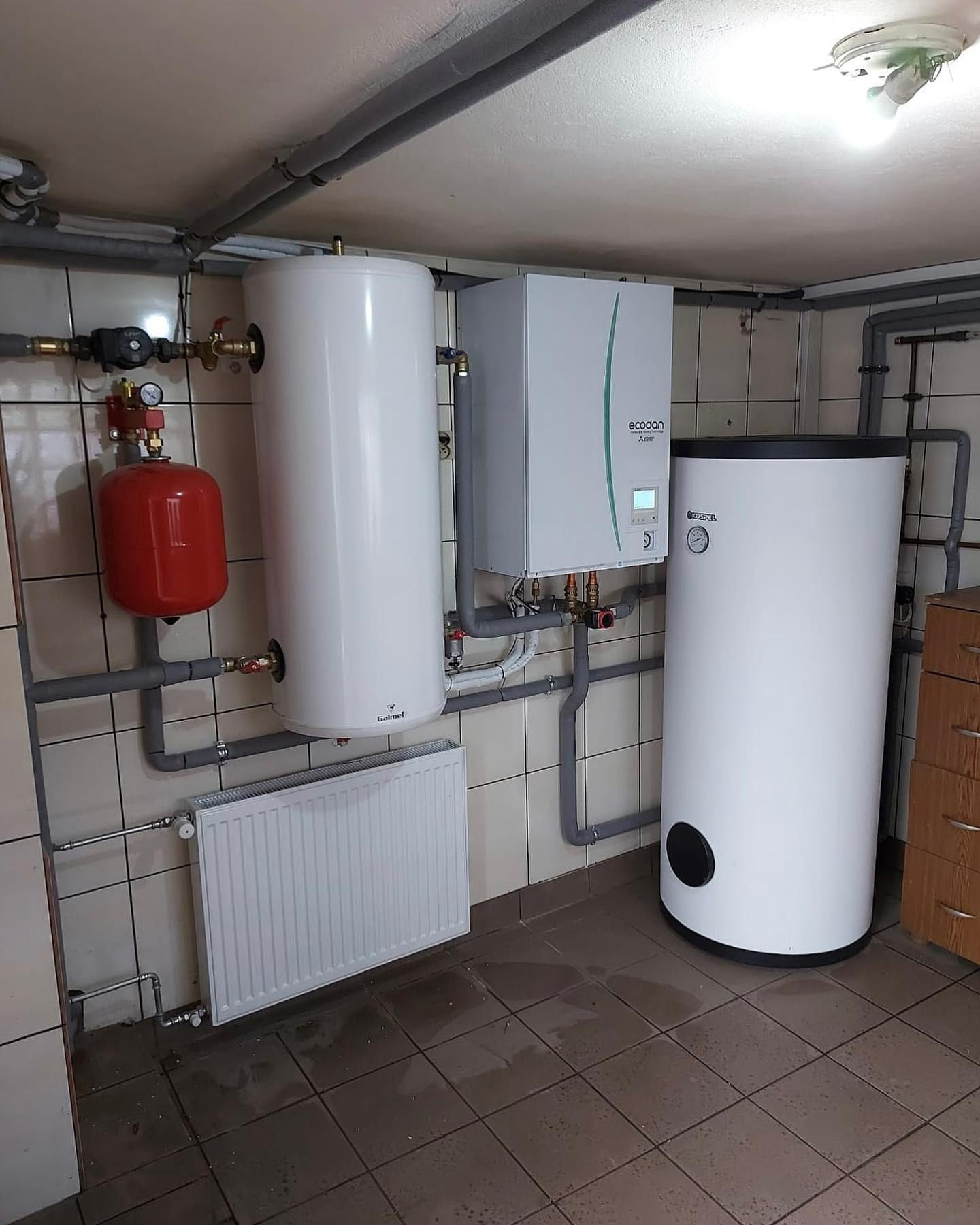 Zestaw urządzeń grzewczych w kotłowni: zasobnik ciepłej wody Galmet, jednostka zewnętrzna Ecodan Mitsubishi Electric, czerwony zbiornik przeponowy, grzejnik panelowy, instalacja rurowa w szarym...