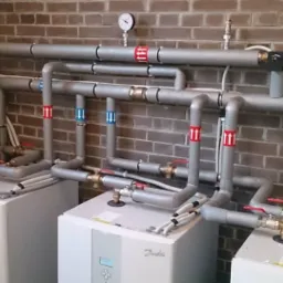 Kaskada trzech gruntowych pomp ciepła Danfoss w kotłowni, z widocznymi rurami, zaworami, manometrem i ceglaną ścianą w tle.