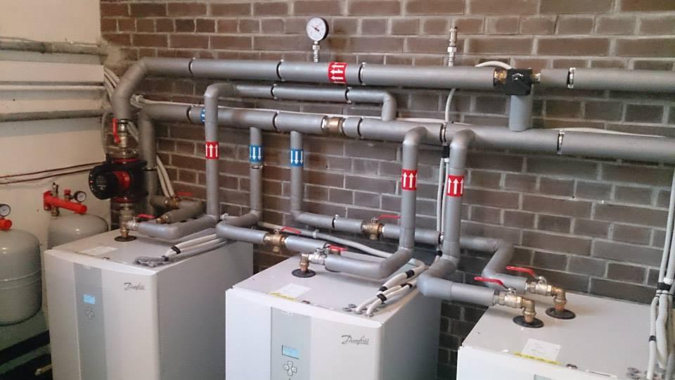 Kaskada trzech gruntowych pomp ciepła Danfoss w kotłowni, z widocznymi rurami, zaworami, manometrem i ceglaną ścianą w tle.