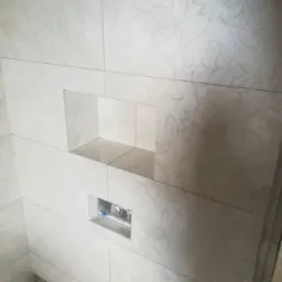 Wykończona ściana w łazience z jasnych, kwadratowych płytek z delikatnym, kwiatowym wzorem, wnęka na kosmetyki i otwór na instalację sanitarną.