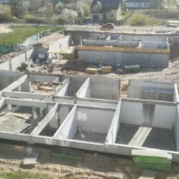 Wykop pod budowę domu jednorodzinnego z widocznymi fundamentami i ścianami działowymi, częściowo w szalunkach, na tle zabudowy mieszkalnej i zieleni.