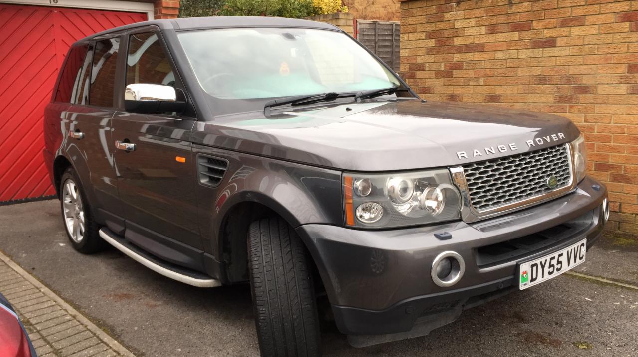 Szary Range Rover Sport zaparkowany przed czerwonym garażem i ceglanym murem, widoczna tablica rejestracyjna DY55 VVC.