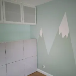 Miętowa ściana z ręcznie malowanym motywem g&oacute;rskim w odcieniach szarości i bieli, uzupełniona białymi szafkami podwieszanymi i stojącymi.