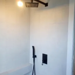 Minimalistyczna łazienka w trakcie wykańczania: białe płytki, czarna armatura prysznicowa (deszczownica i słuchawka), surowa żarówka wisząca na kablu.