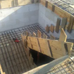 Zbrojenie fundamentów domu z bloczków, widoczne drewniane szalunki i siatka zbrojeniowa, czerwone narzędzie leży na zbrojeniu.