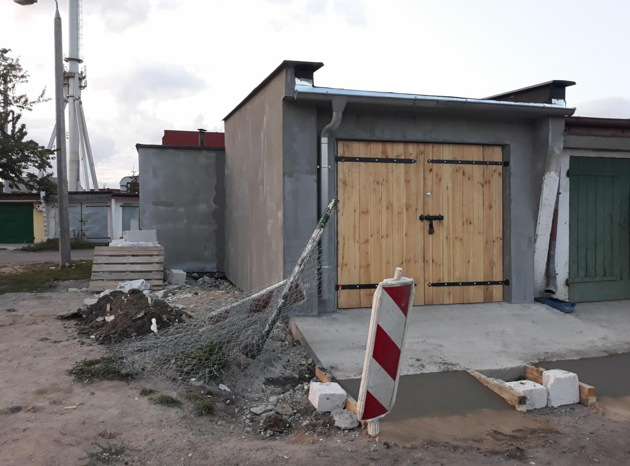 Nowo wybudowany, surowy garaż murowany z drewnianymi wrotami i czarnymi okuciami, widoczna świeża wylewka betonowa przed wjazdem, zabezpieczona siatką i biało-czerwoną barierką, w tle komin...