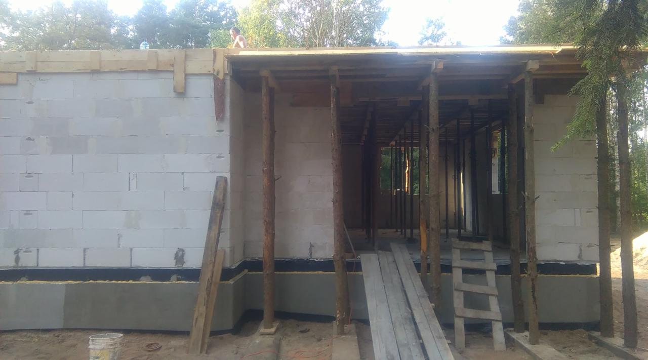 Budowa domu z białych bloczków: surowy stan z drewnianą konstrukcją dachu i kolumnami z bali, widoczne deski i drabina, osoba na dachu w tle.