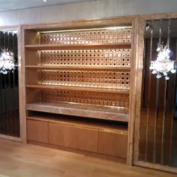 Zabudowa meblowa z półkami i szafkami z jasnego drewna, z marmurowym blatem, podświetlonymi plecami z dekoracyjnymi kwadratami i lustrami po bokach, odbijającymi kryształowy żyrandol.