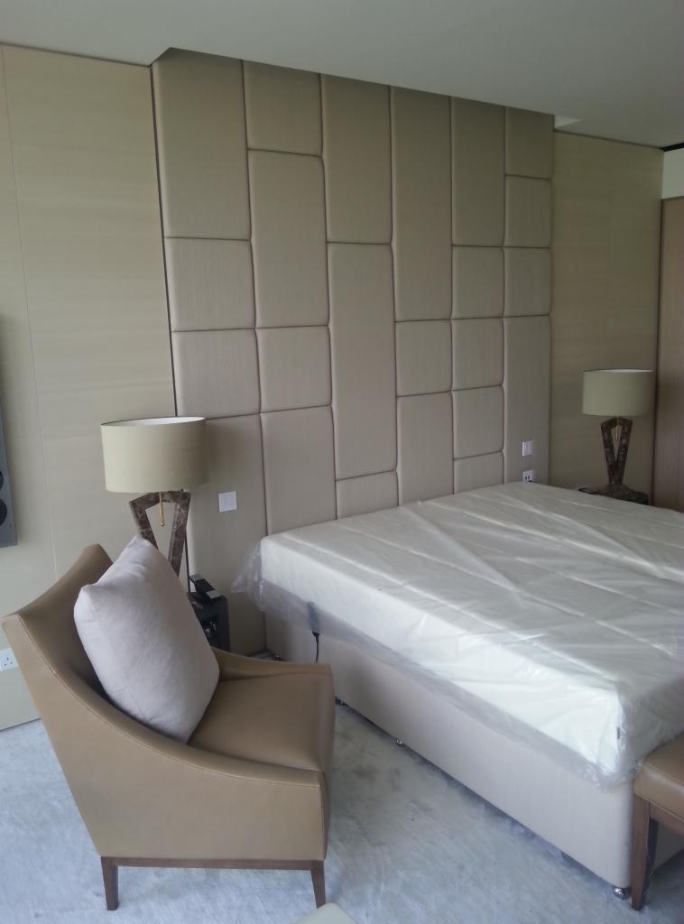 Beżowa sypialnia z tapicerowanym panelem ściennym za łóżkiem, fotel z poduszką i lampy nocne. Łóżko zabezpieczone folią ochronną.
