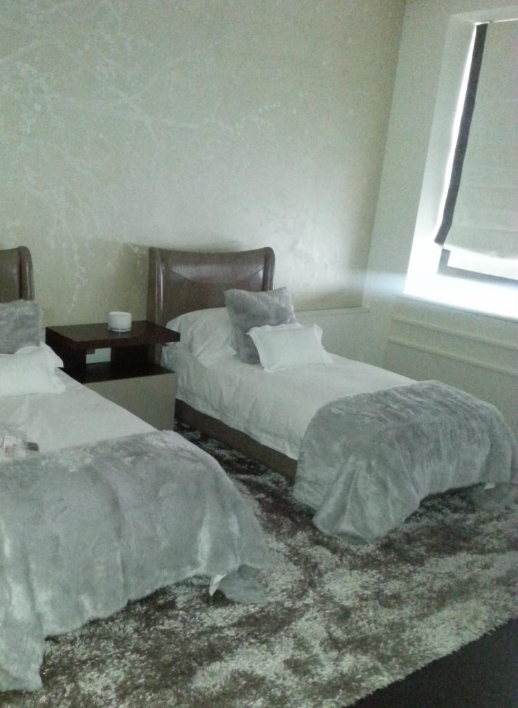 Pokój z dwoma łóżkami pojedynczymi, szary dywan z długim włosiem, ściana z delikatnym wzorem, ciemnobrązowe szafki nocne z białymi akcentami, biała pościel i szare pledy.