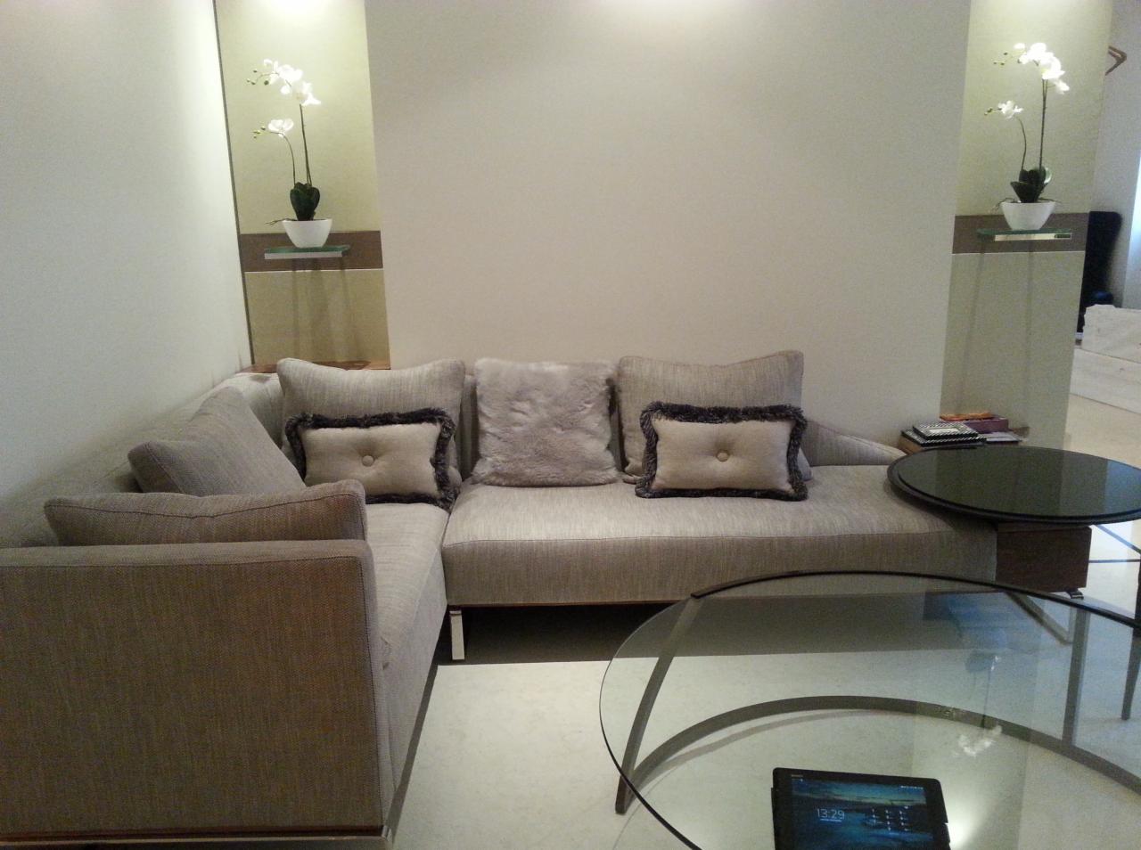 Szary narożnik z poduszkami w salonie, stolik kawowy ze szklanym blatem i tablet na nim, dekoracyjne kwiaty na półkach w tle.