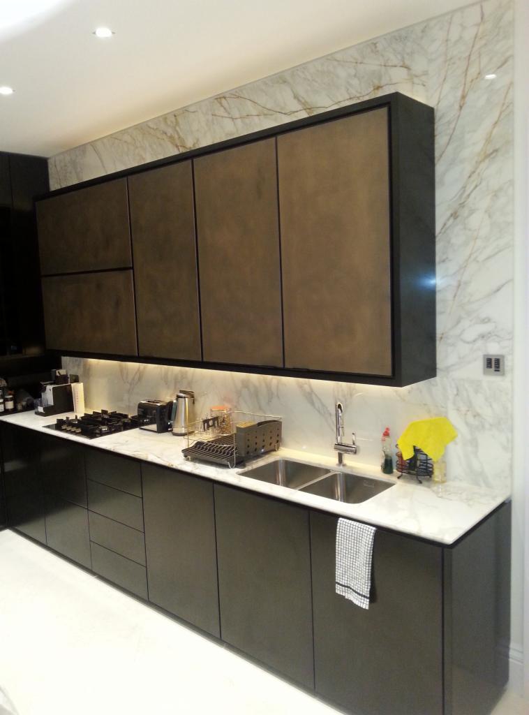 Nowoczesna kuchnia z ciemnymi, matowymi szafkami, marmurowym blatem i ścianą, podświetleniem LED pod szafkami wiszącymi oraz zlewozmywakiem dwukomorowym.