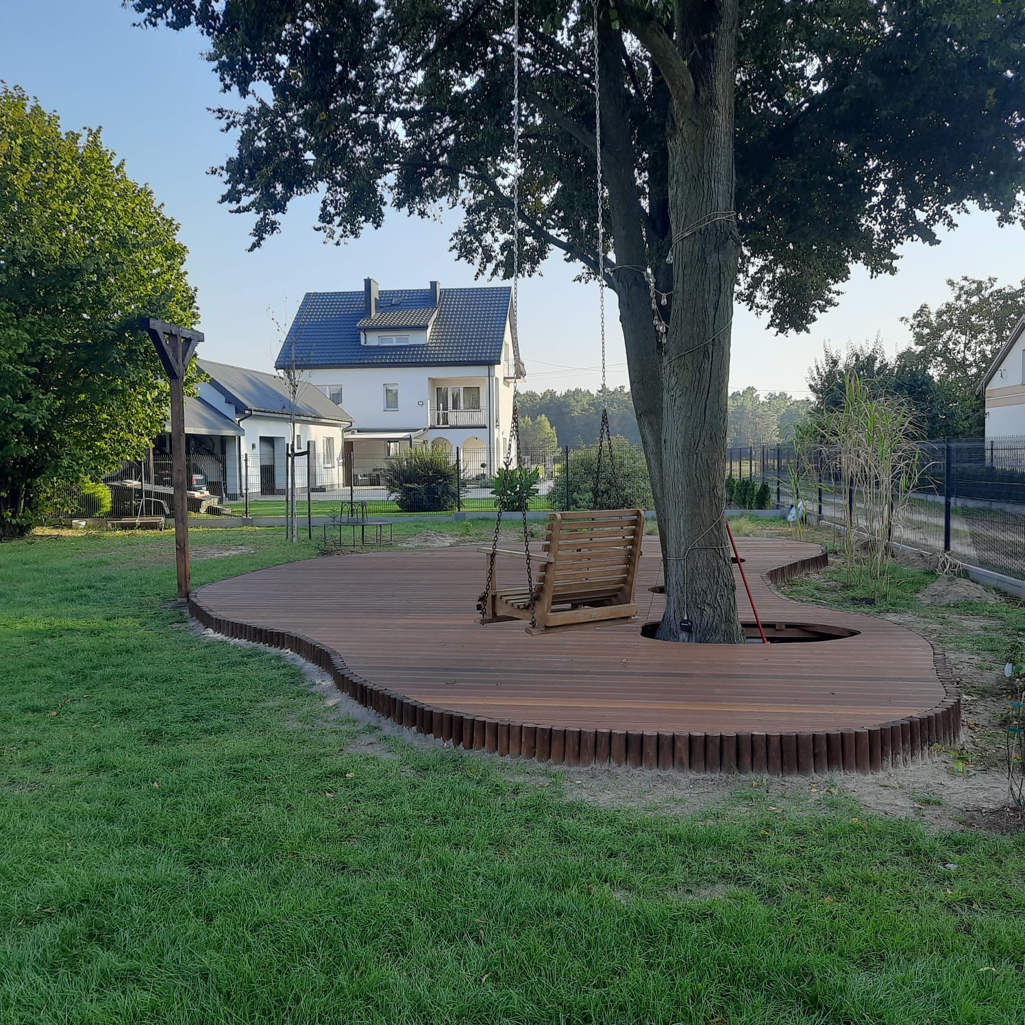Nietypowy taras z drewna otaczający drzewo, z huśtawką zawieszoną na gałęziach, w tle dom jednorodzinny i zieleń ogrodu.