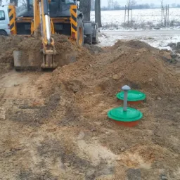 Żółta koparka w trakcie prac ziemnych przy instalacji dwóch zbiorników z zielonymi pokrywami na posesji, w tle widoczny samochód dostawczy i niebieska toaleta przenośna.