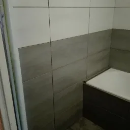 Wykończona łazienka z dwukolorowymi płytkami ceramicznymi ułożonymi do połowy wysokości ściany, widoczny fragment wanny z obudową w ciemnym kolorze.