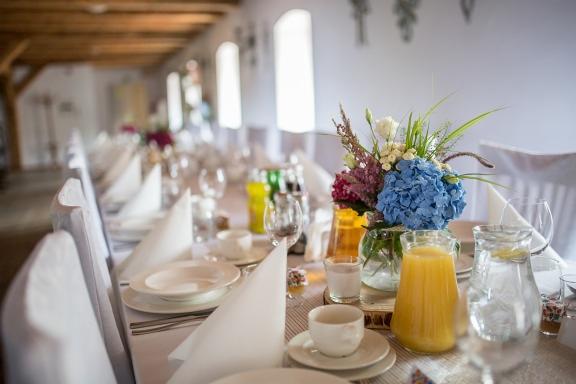 Długi stół weselny z białymi nakryciami, szklankami, filiżankami, dzbankami z sokiem pomarańczowym i wodą oraz bukietem kwiatów z niebieską hortensją w szklanym wazonie na drewnianej podstawce.