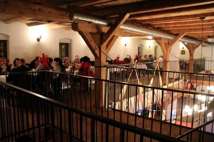 Wnętrze sali z drewnianą konstrukcją stropu, widok z antresoli na grupę osób siedzących przy stołach, część z nich w świątecznych nakryciach głowy, dekoracje na stołach, na dolnym poziomie widoczne...