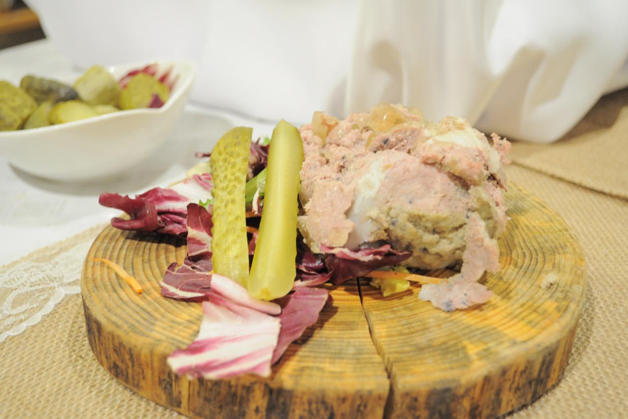 Przystawka z pasztetem, sałatą i ogórkami kiszonymi podana na drewnianej desce, w tle miseczka z ogórkami konserwowymi oraz biała serweta.