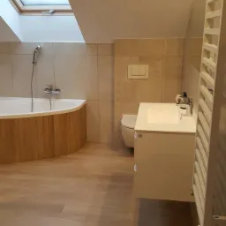 Jasna łazienka na poddaszu z oknem dachowym, beżowe płytki ścienne i podłogowe imitujące drewno, wanna z obudową z drewnianych paneli, biała szafka umywalkowa.