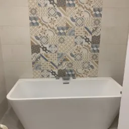 Nowoczesna biała wanna ustawiona pod ścianą z dekoracyjnymi płytkami w odcieniach beżu, brązu i szarości, widoczne baterie wannowe.