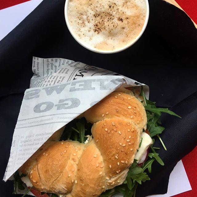 Bajgiel z rukolą i serem, zawinięty w papier śniadaniowy z nadrukiem gazetowym, obok filiżanka cappuccino z cynamonem na czarnej serwetce, ujęcie z góry.