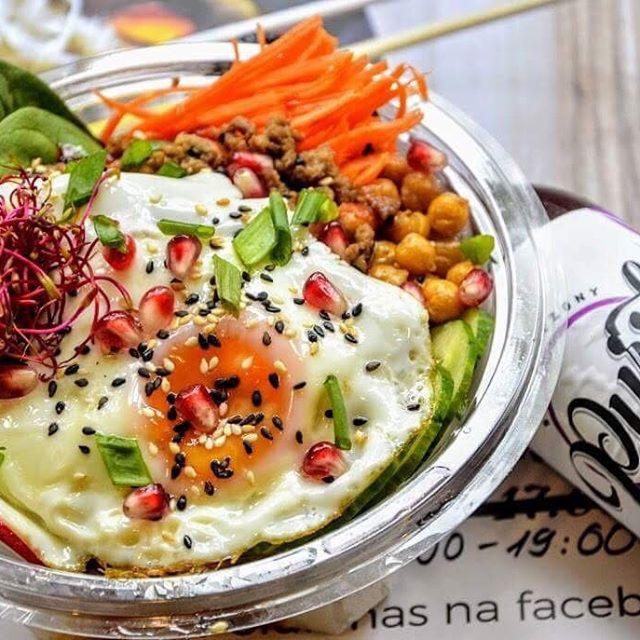 Bibimbap w przezroczystej misce z jajkiem sadzonym, nasionami sezamu, granatem, marchewką julienne i innymi dodatkami, obok napoju z widocznym fragmentem etykiety i grafikiem godzin otwarcia na tle.