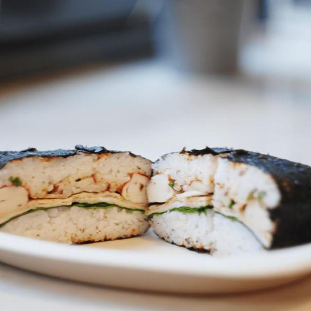 Przekrojone onigirazu na białym talerzu, wypełnione ryżem, jajkiem, zielonymi warzywami i krabem, owinięte w nori.