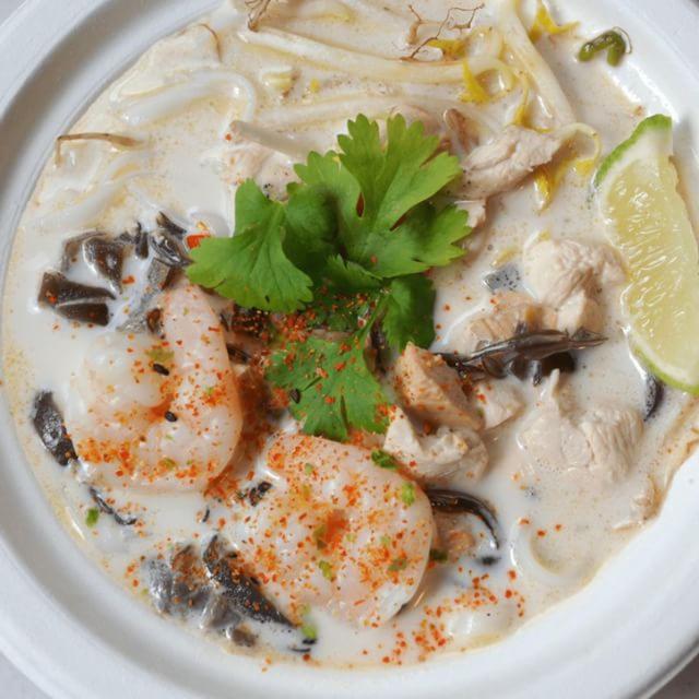 Porcja tajskiej zupy Tom Kha w białej misce, z krewetkami posypanymi przyprawą, kurczakiem, grzybami, świeżą kolendrą i ćwiartką limonki.
