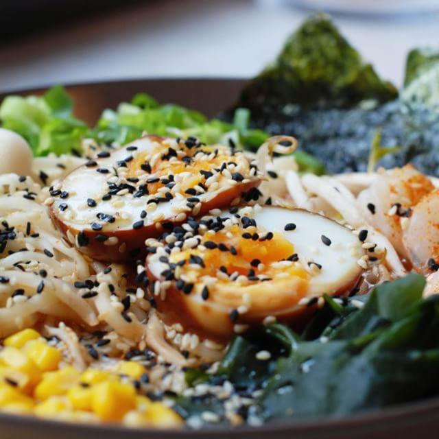 Miseczka z ramenem: dwa przekrojone jajka posypane sezamem, kukurydza, wodorosty nori i wakame, szczypiorek i inne warzywa na tle ciemnej miski.