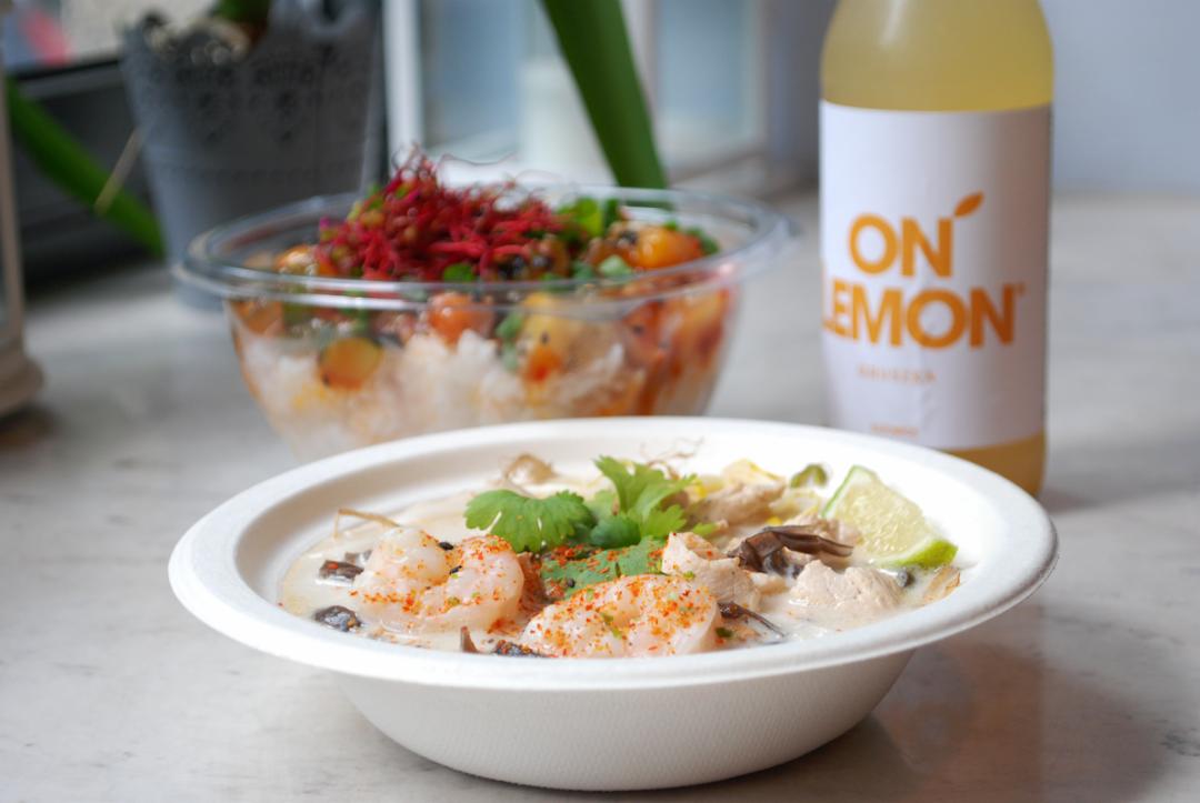 Tajska zupa kokosowa Tom Kha z krewetkami i grzybami w papierowej misce, w tle sałatka ryżowa w przezroczystym pojemniku i butelka lemoniady.