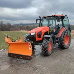 Pomarańczowy traktor Kubota z zamontowanym pługiem do śniegu stoi na błotnistym terenie, w tle pole i pochmurne niebo.