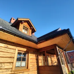 Fragment drewnianego domu z ciemnym dachem, widok z dołu ukazujący detale konstrukcji, okna z firankami i komin z cegły na tle błękitnego nieba.