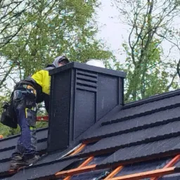 Dekarz w Rychwałdzie podczas montażu komina na dachu pokrytym czarną dachówką bitumiczną, widoczna więźba dachowa i narzędzia pracy.