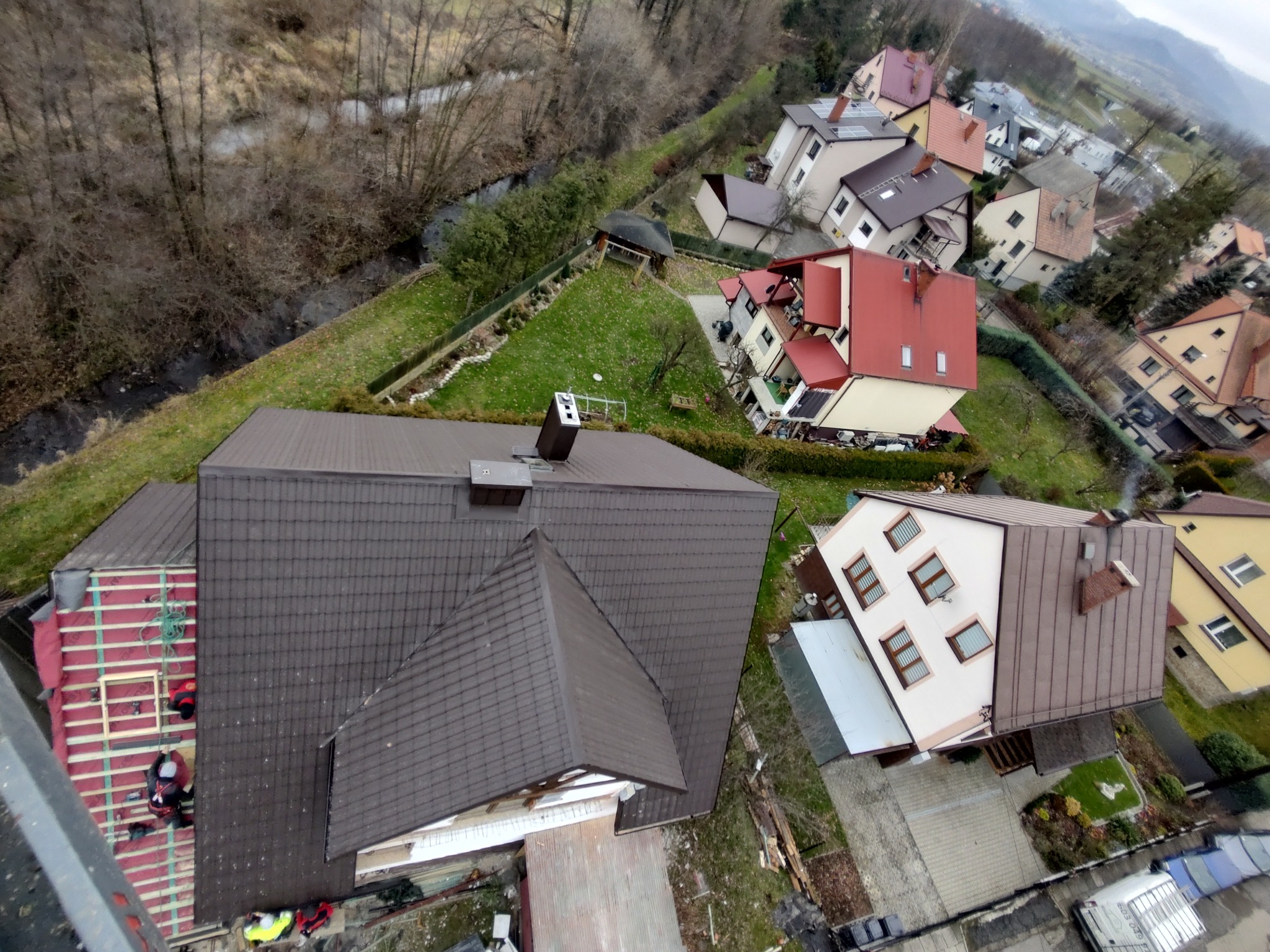 Widok z góry na dom w trakcie budowy: część dachu pokryta brązową dachówką, druga część z widoczną konstrukcją drewnianą i ekipą montującą, w tle zabudowa jednorodzinna i rzeka.