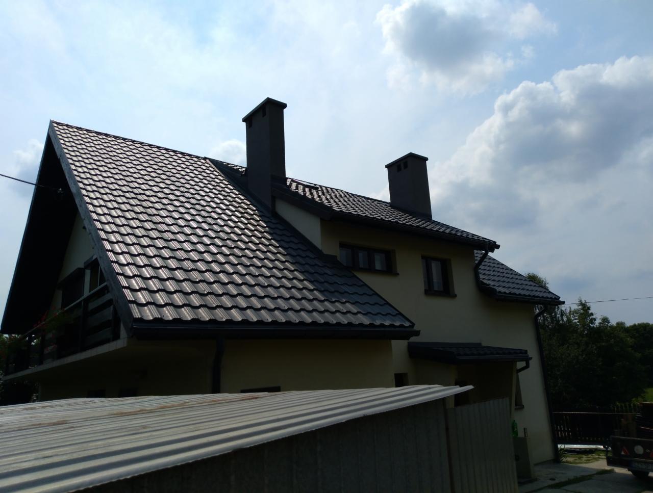 Dom jednorodzinny w Rychwałdzie z ciemną dachówką, widok z dołu na dach i komin na tle pochmurnego nieba.