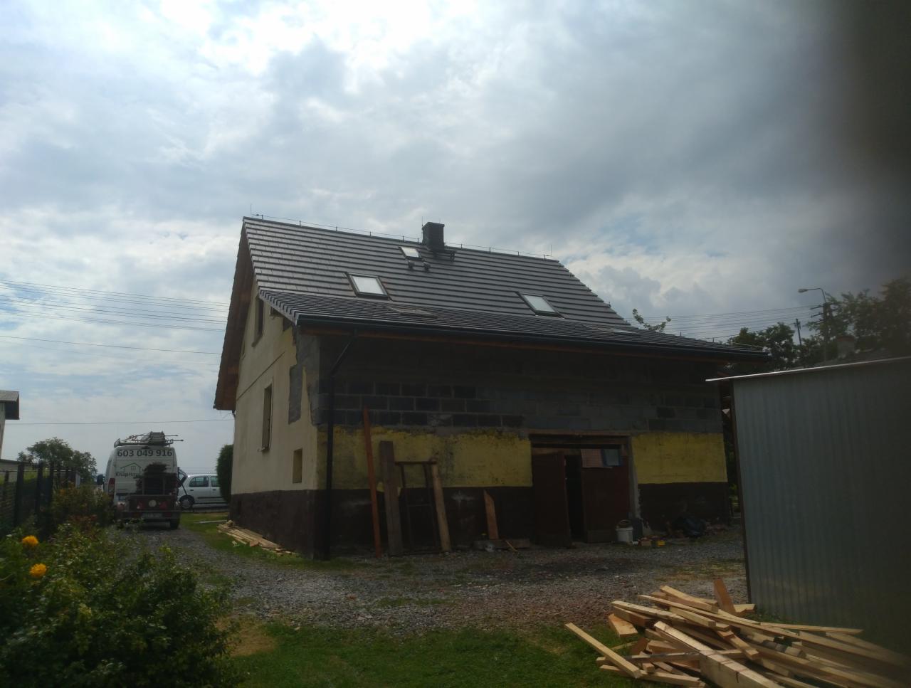 Budynek gospodarczy w trakcie remontu z nowym, grafitowym pokryciem dachowym z oknami połaciowymi; ściany częściowo otynkowane, częściowo z pustaków, w tle zaparkowany samochód dostawczy.