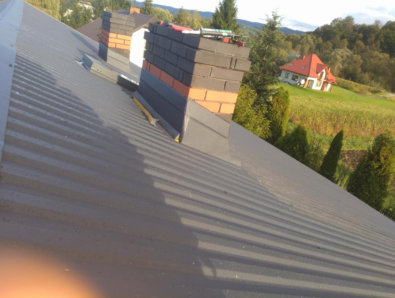 Szary blaszany dach z widocznymi kroplami wody, komin z cegły w dwóch odcieniach oraz krajobraz z domem w tle.