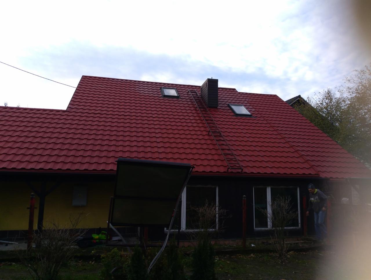 Czerwony dach z blachodachówki, widoczne okna dachowe i komin. Przystawiona drabina do komina. Fragment budynku z żółtą elewacją po lewej stronie i ciemną elewacją po prawej.
