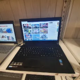 Czarny laptop Lenovo z otwartą przeglądarką internetową wyświetlającą portal informacyjny, stojący na białym regale obok innego laptopa, w tle drewniana ściana i metalowy regał.
