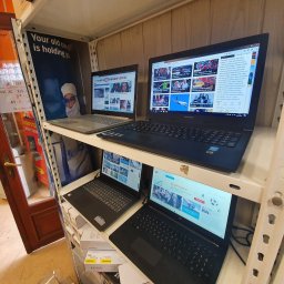 VERTES - serwis komputerów i laptopów w Otwocku - Półka w sklepie komputerowym z czterema używanymi laptopami różnych marek (Lenovo, HP) z wyświetlonymi stronami internetowymi z wiadomościami, obok reklama serwisu komputerowego.