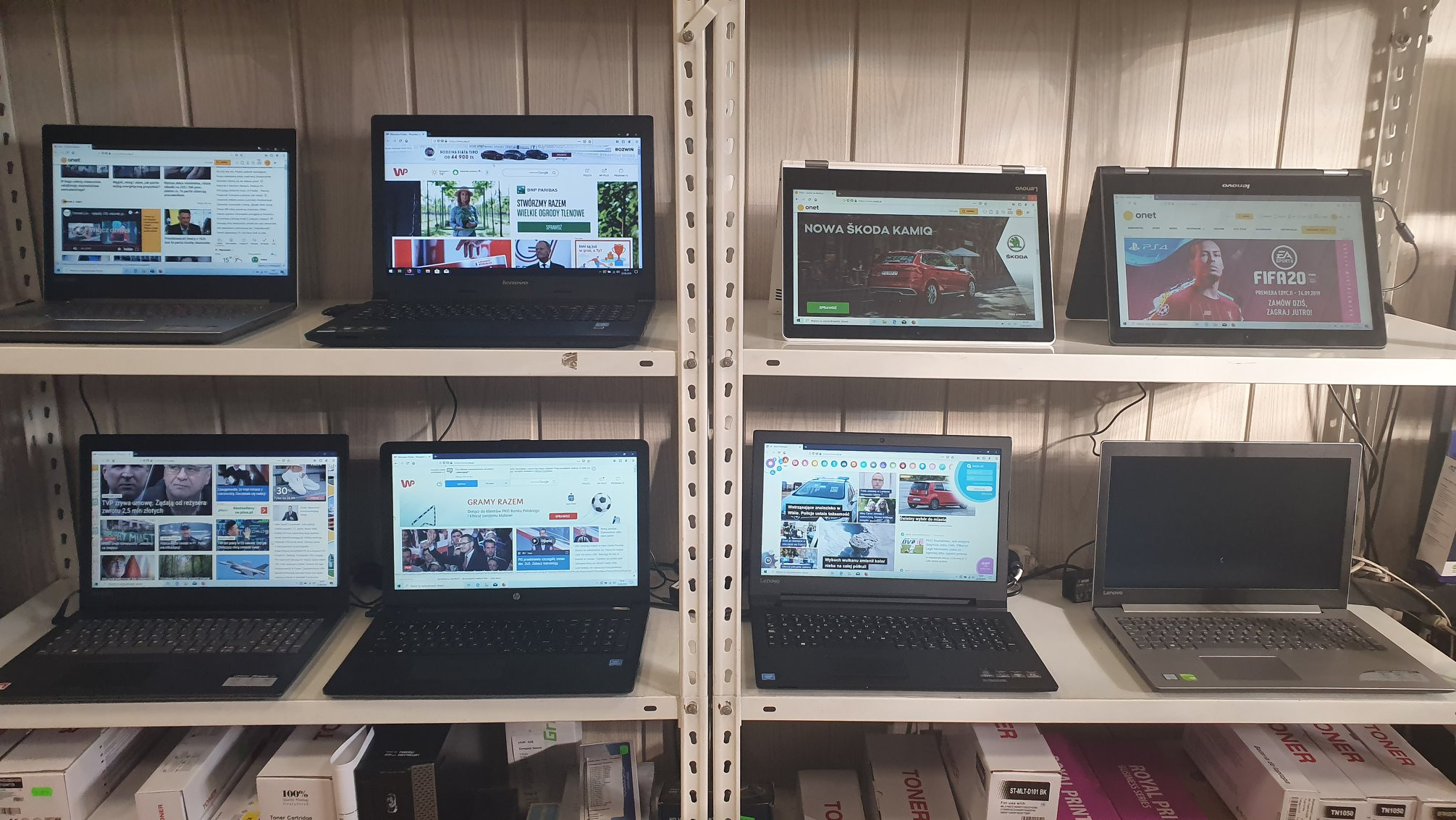 Półka w sklepie z elektroniką, na której ustawione są laptopy różnych marek, w tym Lenovo i HP, z wyświetlanymi stronami internetowymi i reklamami; widoczne kartony z tonerami pod spodem.