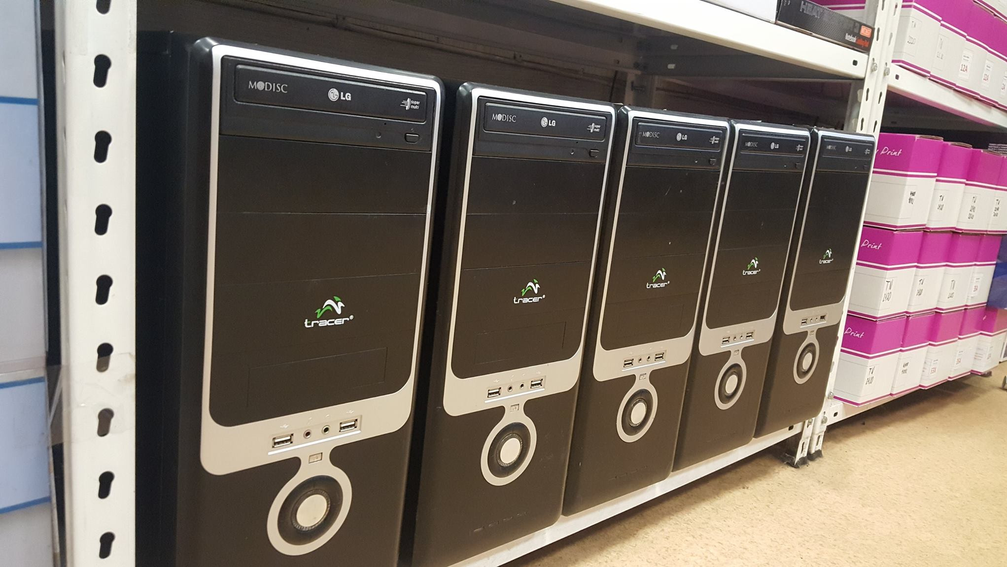 Rząd czarnych obudów komputerowych marki Tracer i LG na półce magazynowej, widoczne pudełka z nadrukiem w tle.