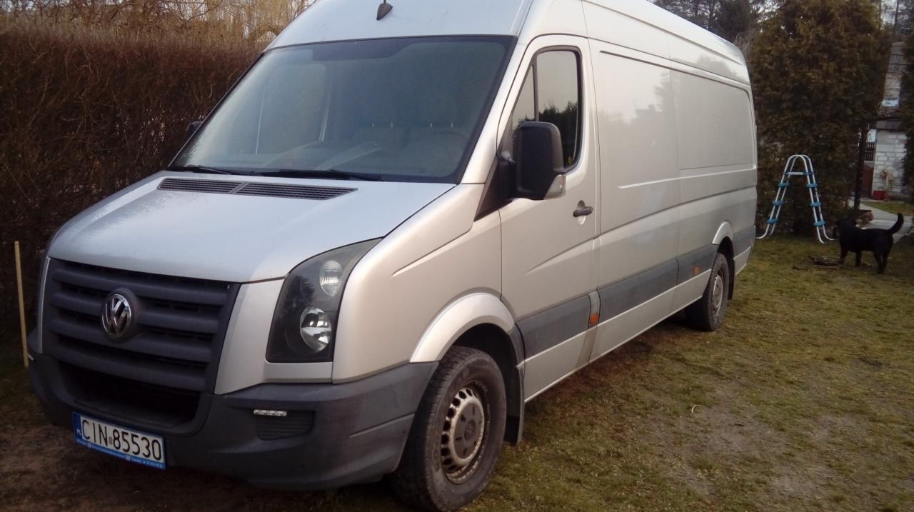 Srebrny samochód dostawczy Volkswagen Crafter z polską tablicą rejestracyjną CIN 85530, zaparkowany na trawniku przed domem z drabinką i psem w tle.