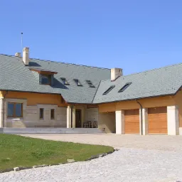 Rozległy dom jednorodzinny z szarym dachem pokrytym dachówką bitumiczną, widoczne okna dachowe, elewacja w odcieniach beżu i piaskowca, garaż z drewnianymi bramami.