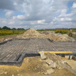Przygotowany szalunek i zbrojenie płyty fundamentowej na budowie domu jednorodzinnego, widoczne zabezpieczenie folią kubełkową, poziomica i fragmenty gruzu, w tle hałda ziemi i łąka z kwitnącymi...