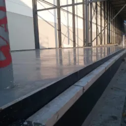 Świeżo wylana, gładka posadzka wewnątrz hali w budowie, widoczny szalunek i słup z czerwoną farbą.